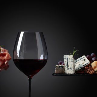 L'immagine raffigura un esempio di abbinamento cibo-vino con formaggi, uva, noci e prosciutto accompagnati da un bicchiere di vino rosso.