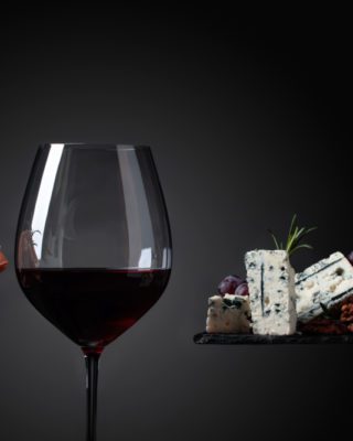 L'immagine raffigura un esempio di abbinamento cibo-vino con formaggi, uva, noci e prosciutto accompagnati da un bicchiere di vino rosso.